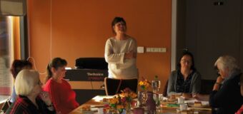 Die Personen sitzen an einem Tisch, gedeckt mit Kaffee und Kuchen, Wasserflaschen und Gläsern und Blumen