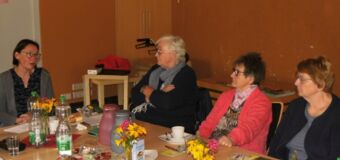 Die Personen sitzen an einem Tisch, gedeckt mit Kaffee und Kuchen, Wasserflaschen und Gläsern und Blumen