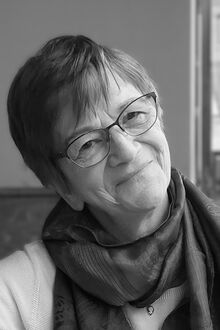 Eva trägt einen Kurzhaarschnitt und eine Brille, hat ein Tuch um den Hals und lächelt in die Kamera