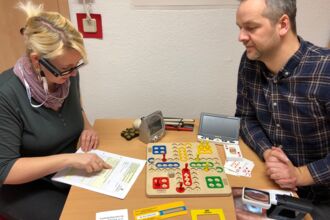 eine Frau hat den Zeigefinger auf einem Formular und versucht mit der Orcam den Inhalt zu lesen, der Berater beobachtet sie. Auf dem Tisch liegen weitere Hilfsmittel