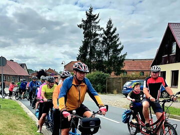 Eine Radgruppe fährt auf einer asphaltierten Dorfstraße. Im Vordergrund ist ein Mann auf einem blauen Tandemrad zu sehen, der den Betrachter anschaut; er trägt ein gelb-blaues Trikot, rosa Handschuhe und einen Helm.