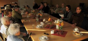 Die Personen sitzen an einem Tisch, gedeckt mit Kaffee und Kuchen, Wasserflaschen und Gläsern und Blumen