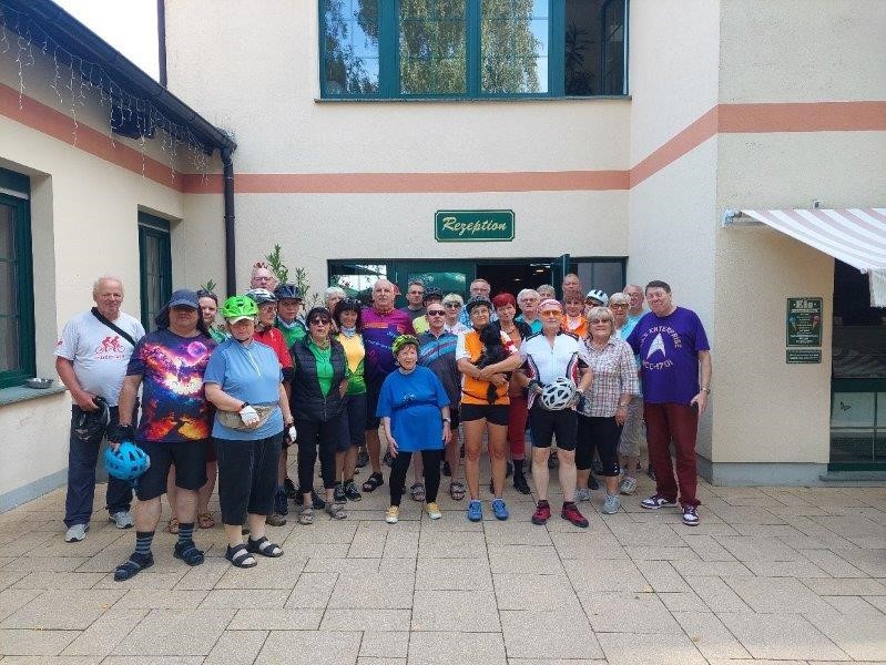 Etwa 25 Personen stehen in Rad- und Freizeitkleidung vor dem Eingang eines Hotels mit dem Schild "Rezeption"